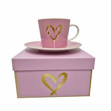 Ružová šálka s podšálkou a zlatým srdcom, balená v ružovej krabičke.